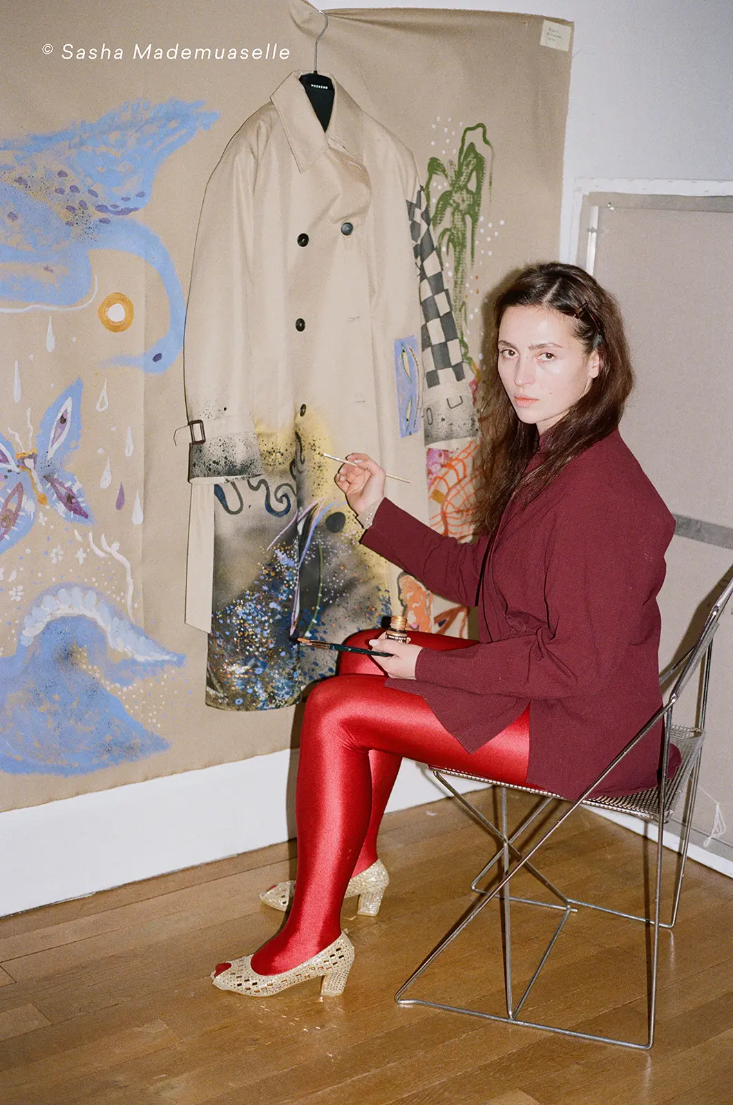 Victoria Kosheleva (the artist) hand-painting the trench coat prototype, seated on a wire chair amidst sketches and blue swan motifs.