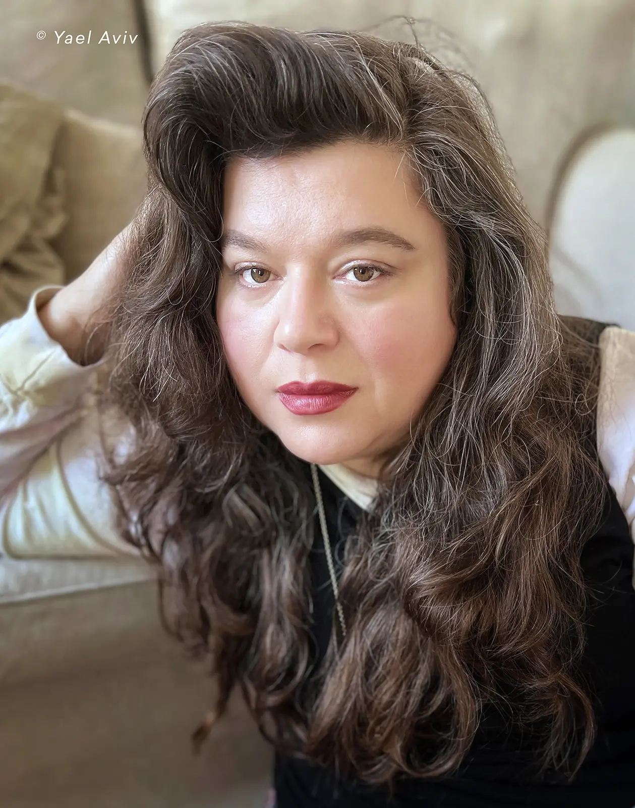 A close-up portrait of a Tai Shani with a calm expression. She has long, wavy dark brown hair with visible gray strands. She is wearing a white shirt and a dark vest, with a subtle necklace.